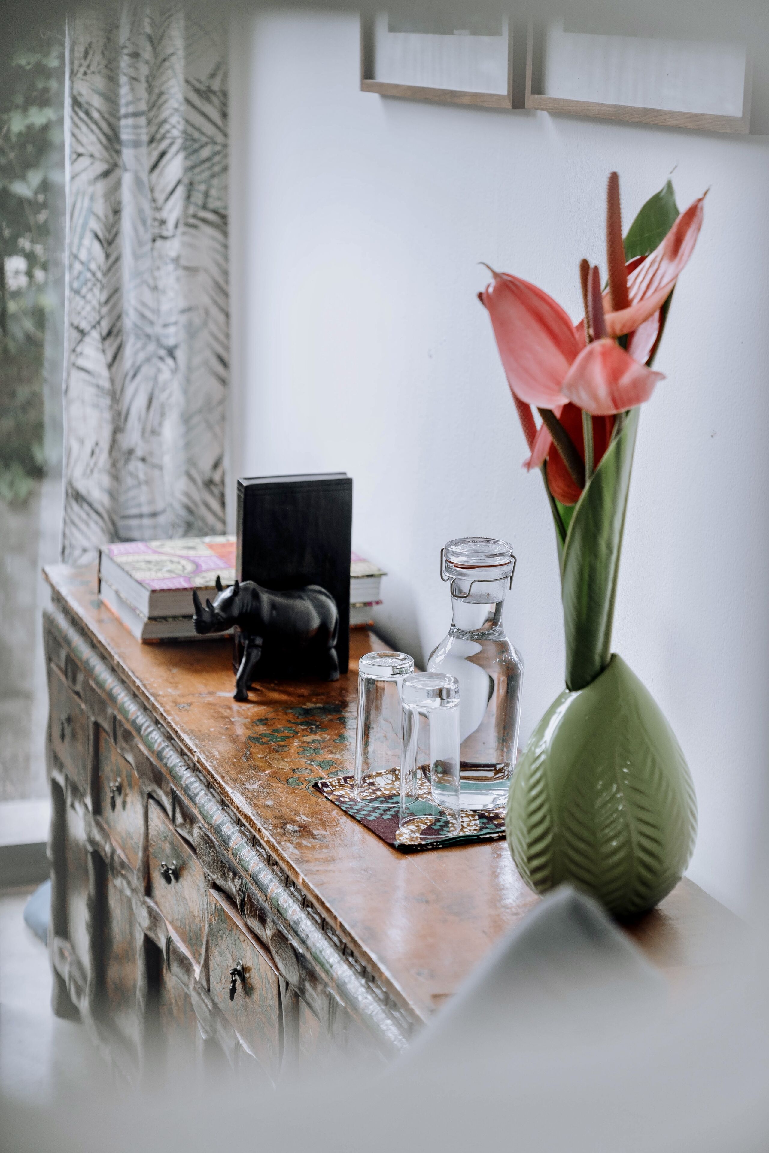 A wooden table with a green vase containing pink flowers, a black figurine, and a few glass bottles.