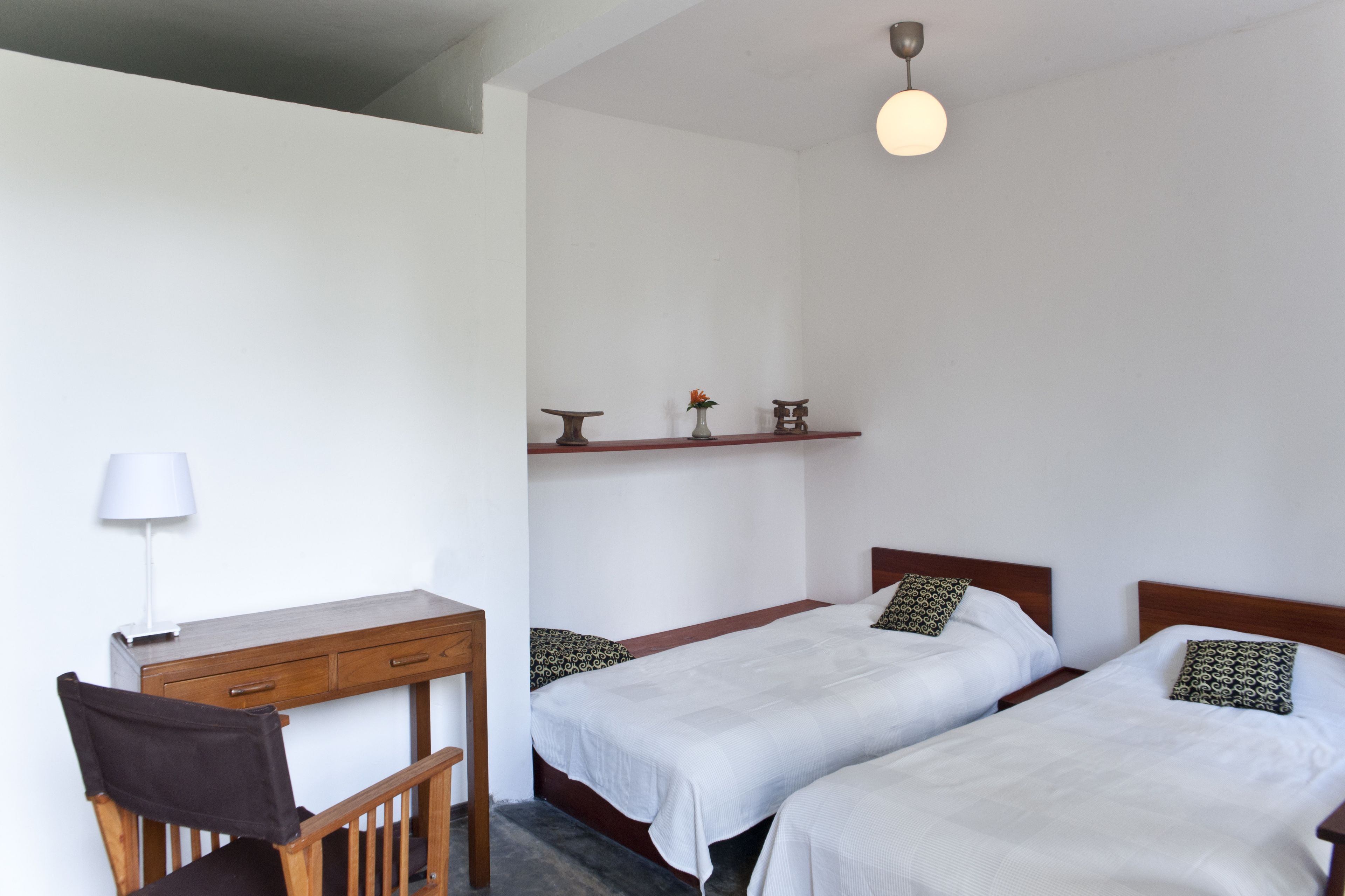 A room with two single beds, a desk with a chair, a lamp, and a shelf with decorative items.