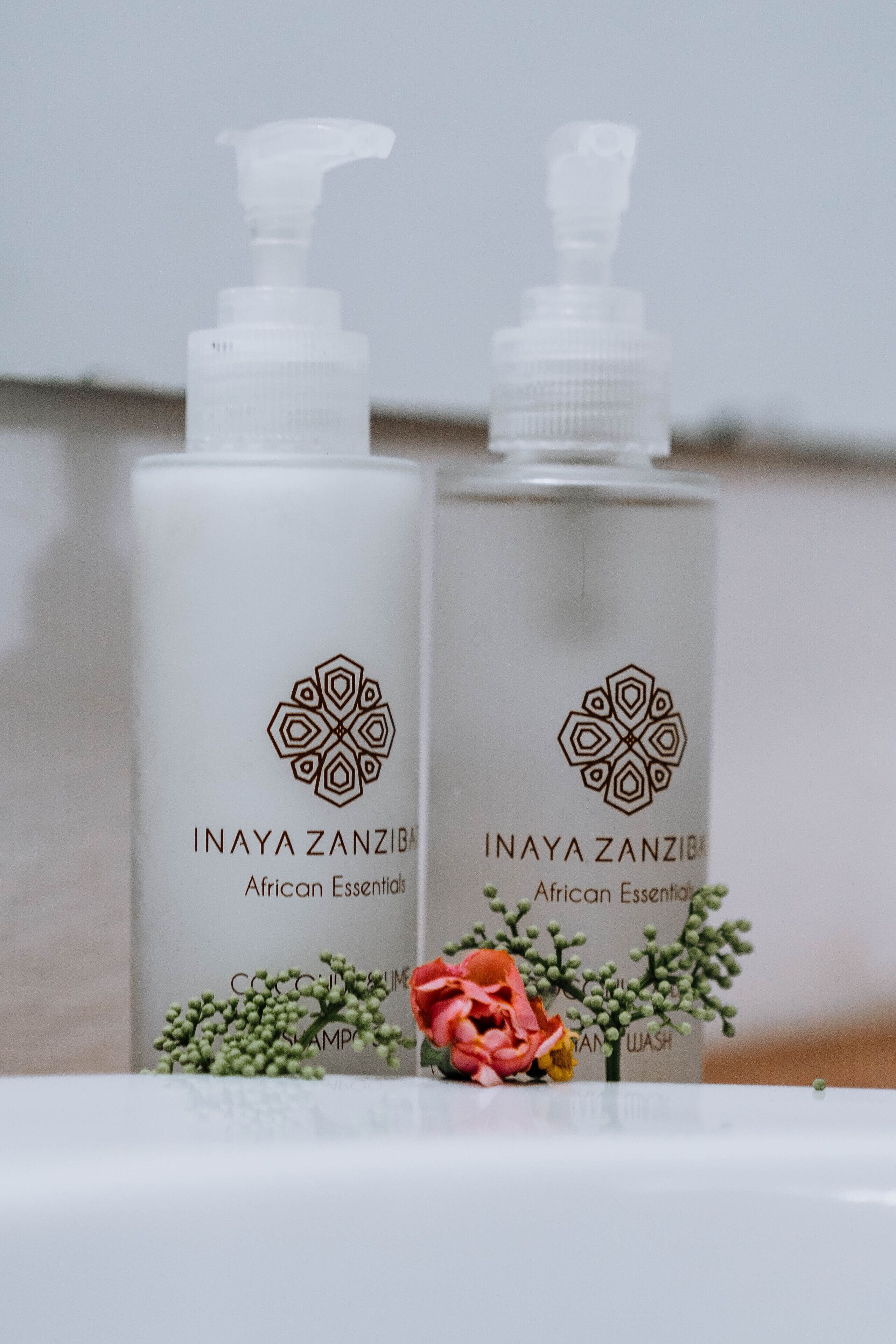 Dos botellas de Inaya Zanzibar African Essentials sobre una superficie blanca, adornadas con flores decorativas.
