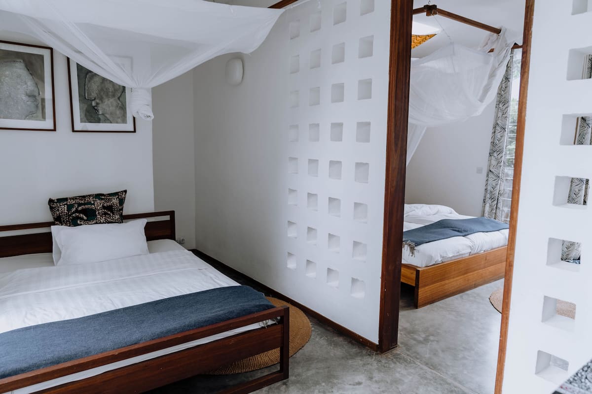 A bedroom with two beds, one with a canopy and one without. There are framed pictures on the wall and a white partition with square cutouts.