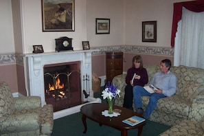 A living room with a fireplace, two sofas, a coffee table with a book and flowers, and framed pictures on the wall. - Lucinda B&B (Latrobe)