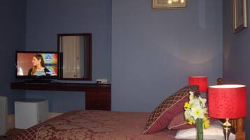 A bedroom with a TV on a stand, a bed with a red and gold patterned bedspread, a nightstand with a red lamp, and a vase with flowers.