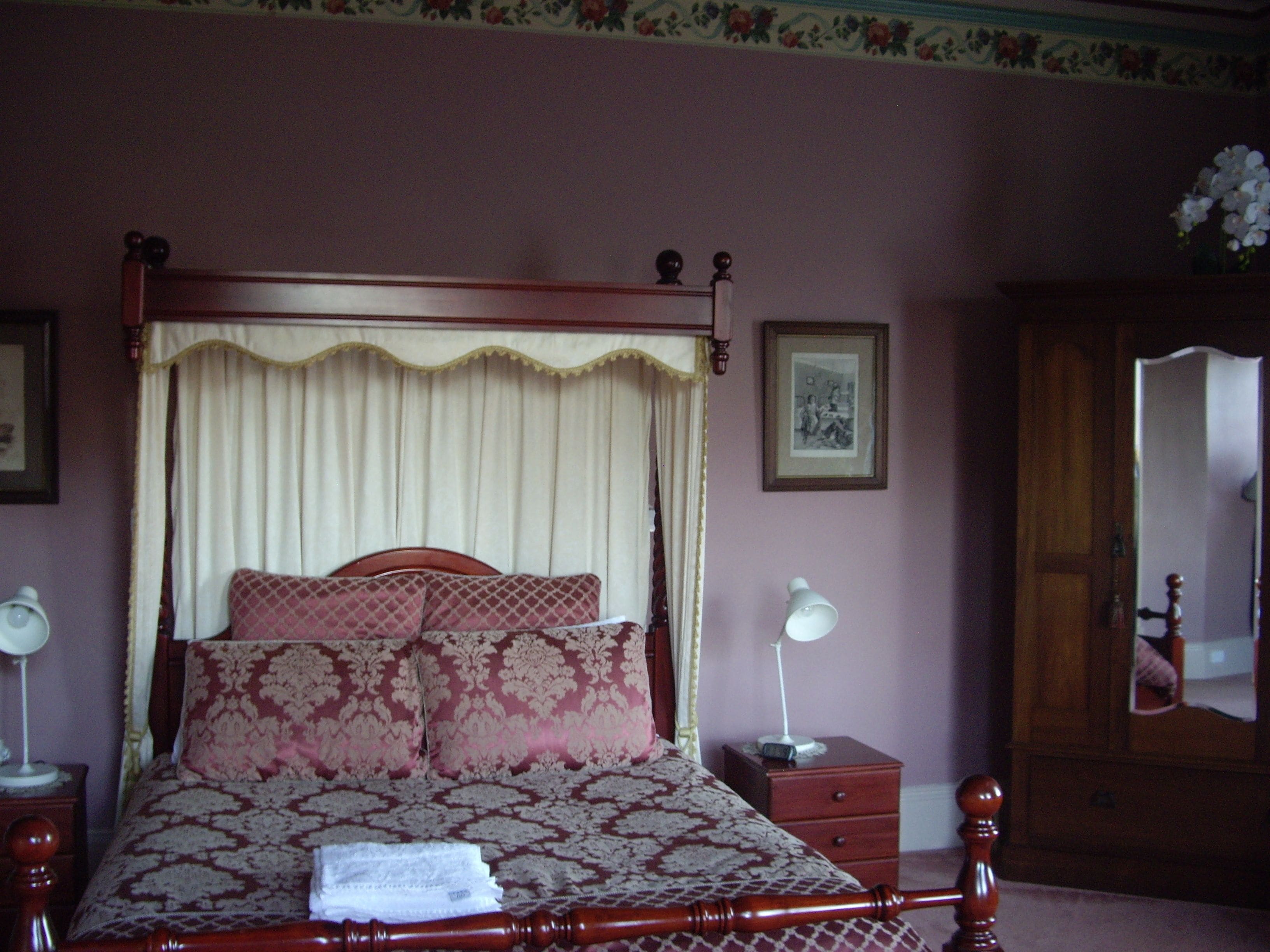 A bedroom with a four-poster bed, a nightstand with a lamp, a dresser, and a mirror.