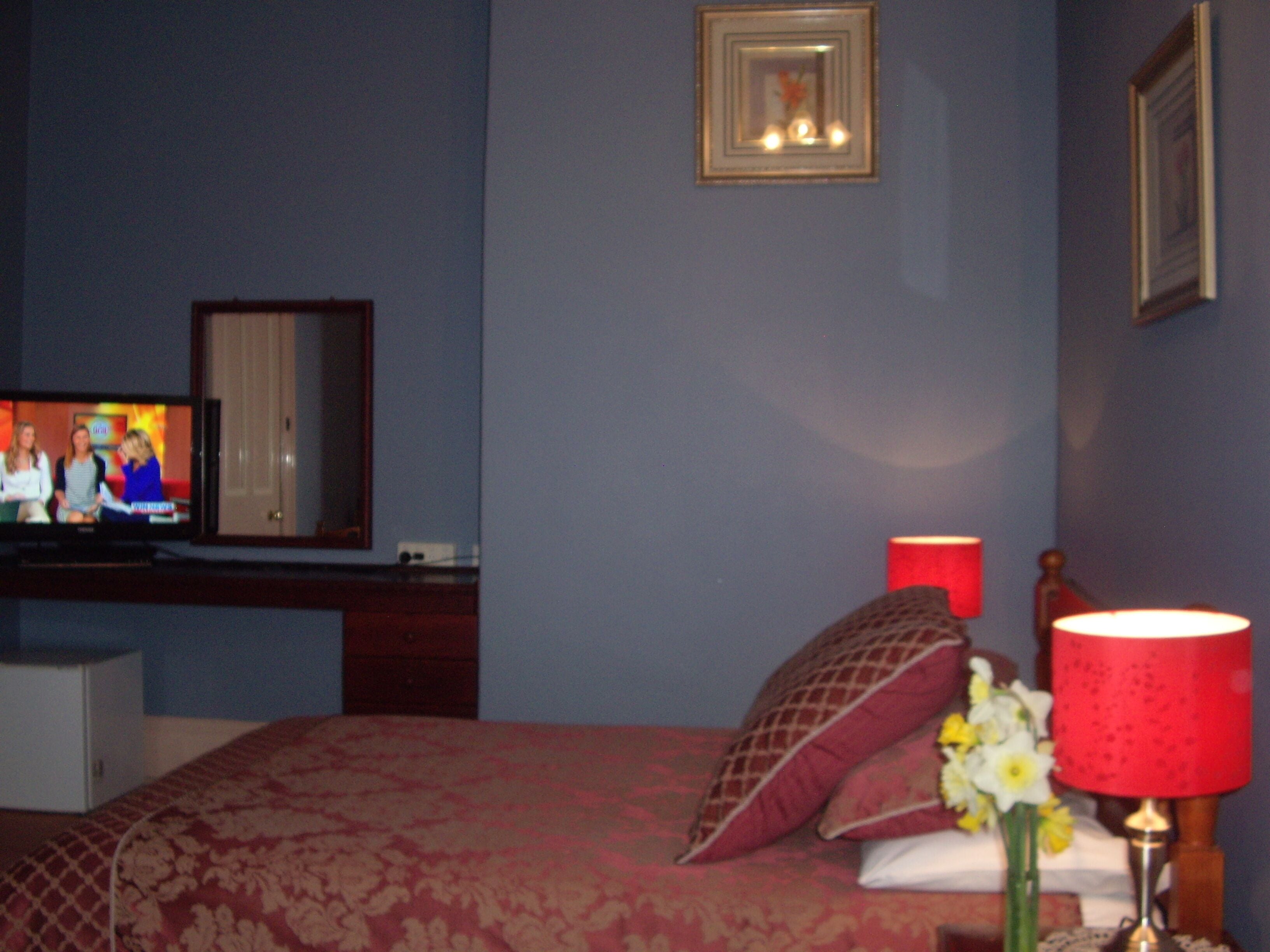 A bedroom with a bed, a television, a mirror, a red lamp, and a framed picture on the wall.