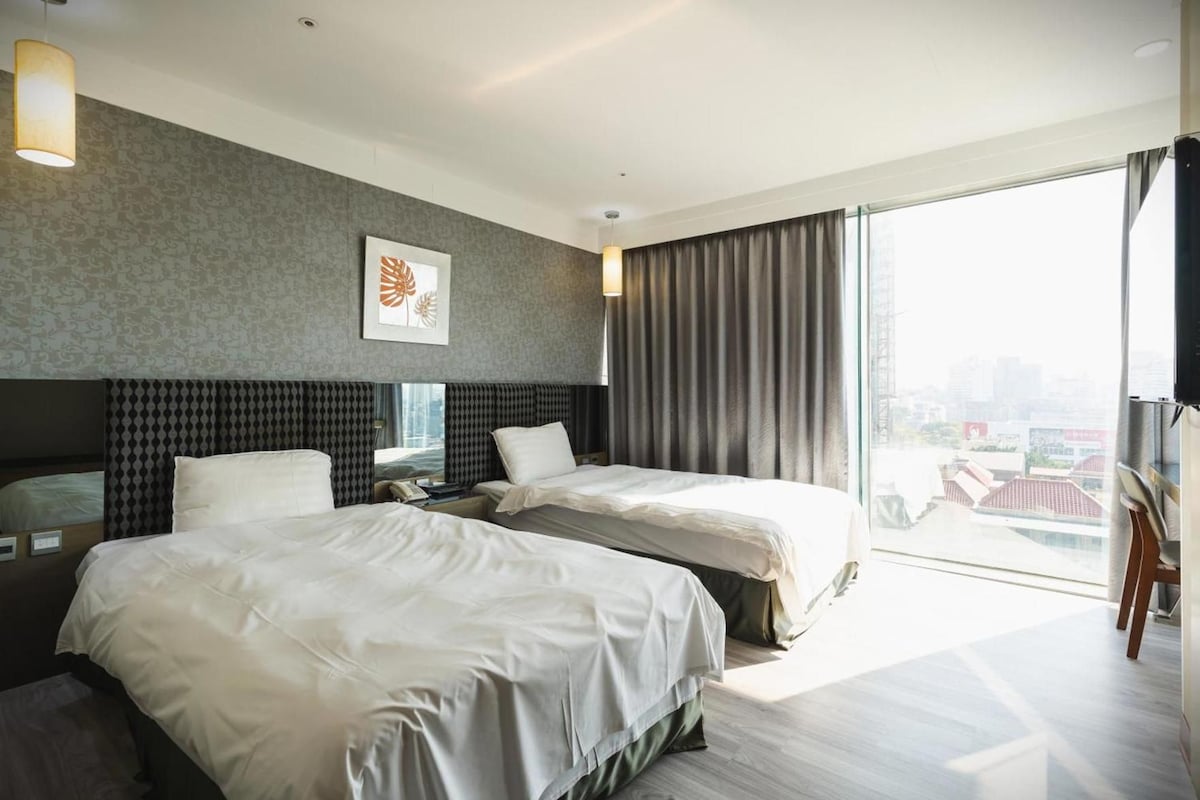 Ein Hotelzimmer mit zwei Betten, einem großen Fenster mit Vorhängen, Blick auf die Stadt und einem gerahmten Kunstwerk an der Wand.