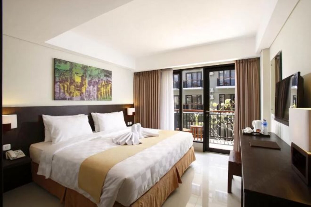 A hotel room with a large bed, a desk, a chair, a TV, and a balcony with a view of the city.