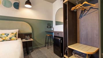 Ett hotellrum med en säng, ett skrivbord, ett kylskåp, en klädstång och en spegel.