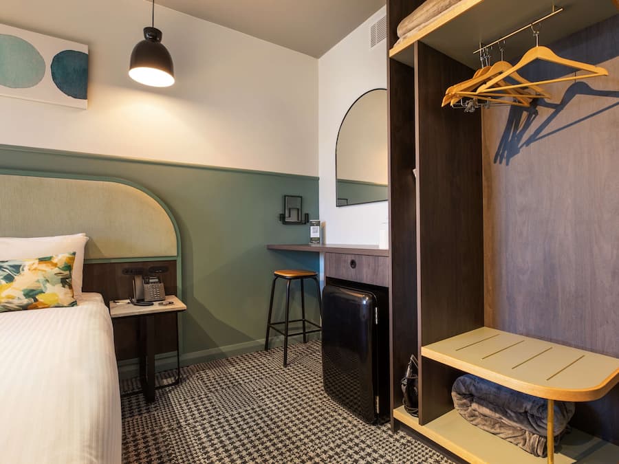 Ett hotellrum med en säng, ett skrivbord, ett kylskåp, en klädstång och en spegel.