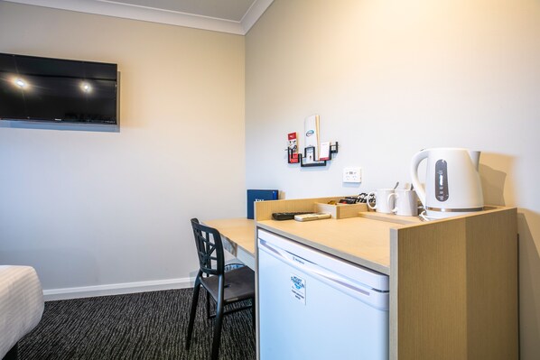 Une chambre d’hôtel de taille réduite, comprenant un lit simple, un petit bureau avec une bouilloire, un réfrigérateur, un téléviseur fixé au mur et une chaise.