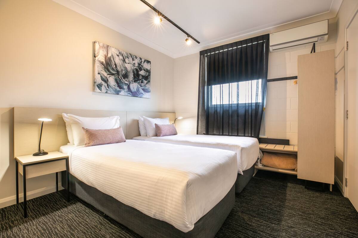 Habitación de hotel con dos camas, un escritorio, un armario y un cuadro abstracto grande en la pared.