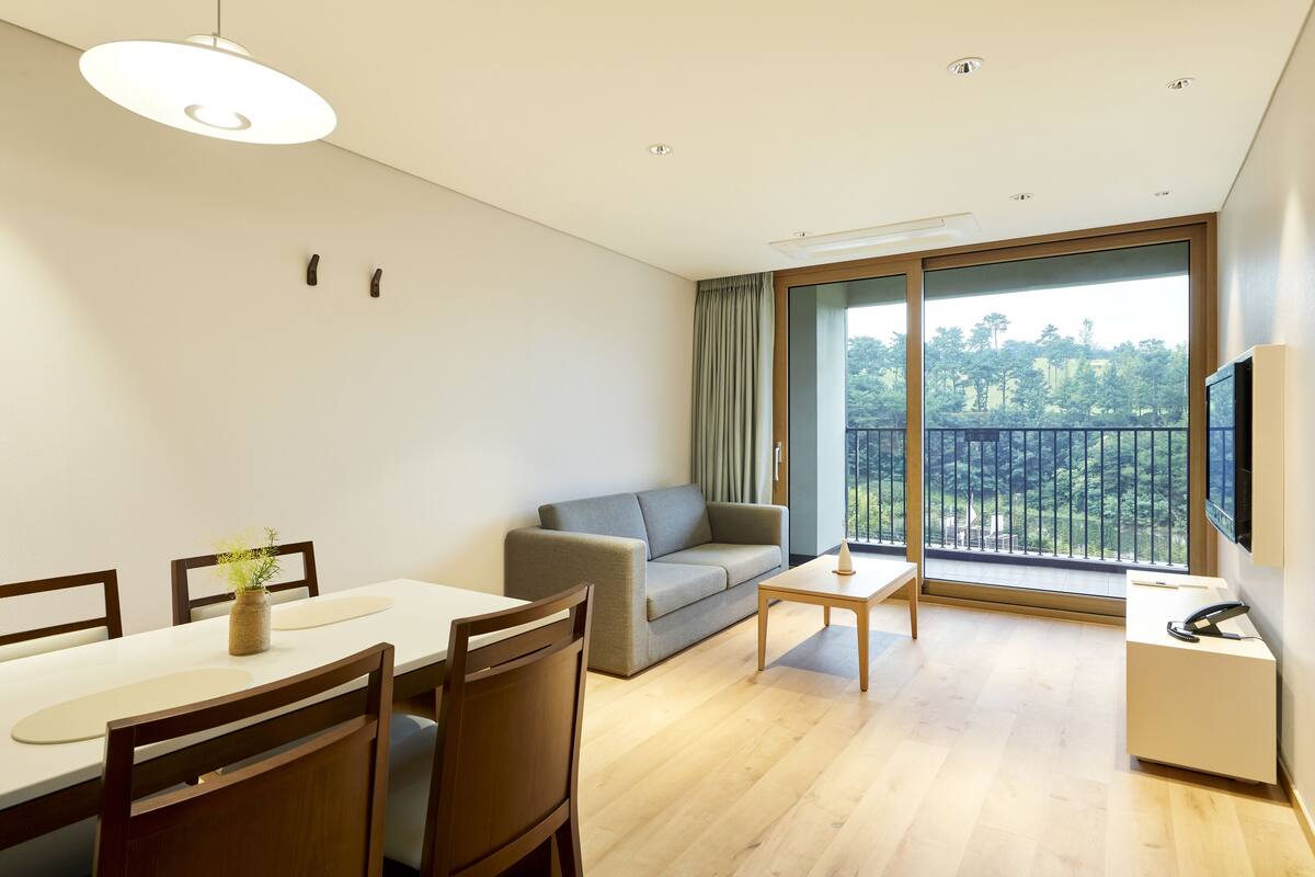 A modern living room with a sofa, dining table, and chairs. There is a TV on the wall and a balcony with a view of trees.