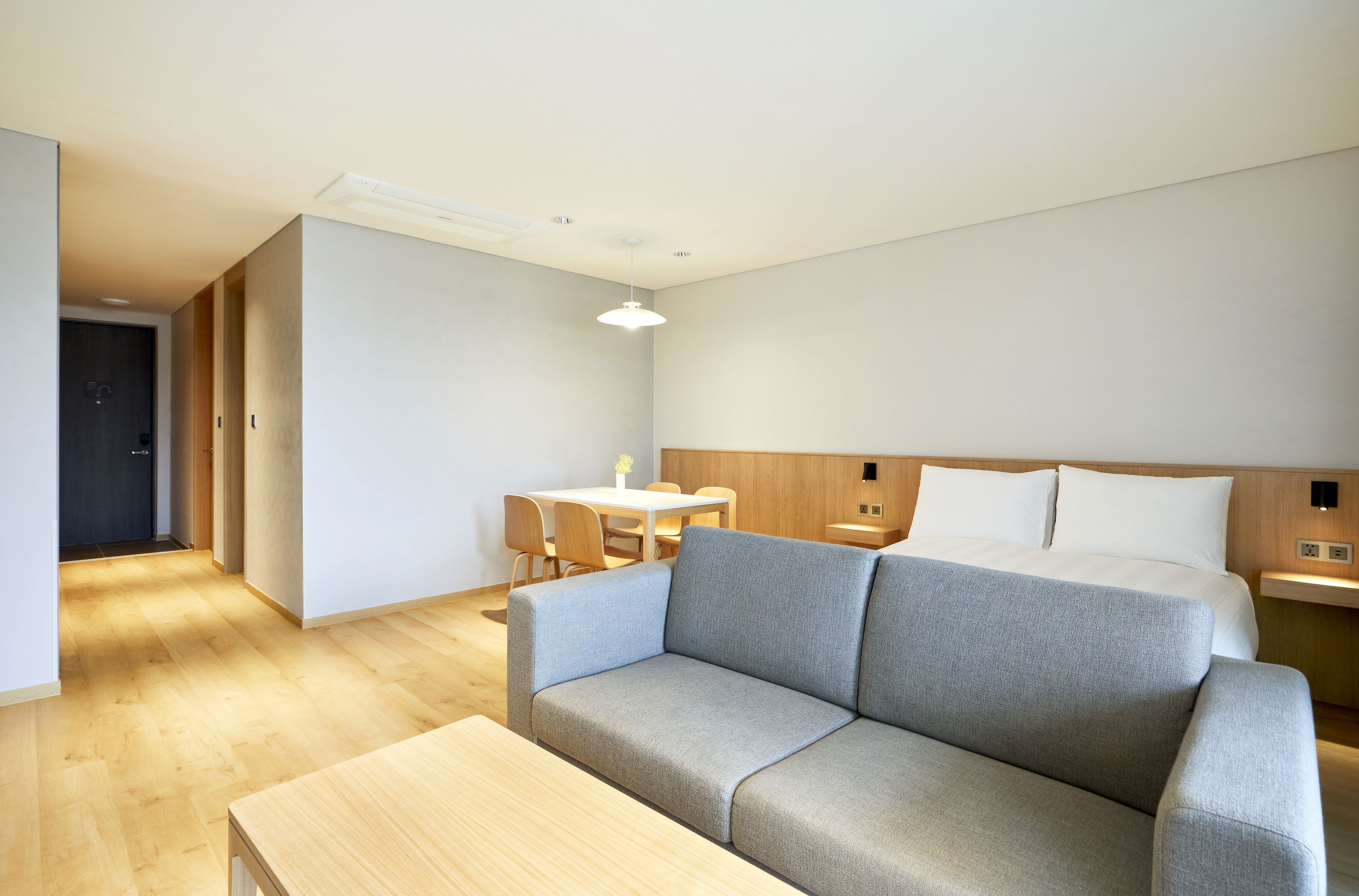 Ein modernes Hotelzimmer mit grauem Sofa, einem Holztisch, einem Bett mit weißer Bettwäsche und einem kleinen Schreibtisch mit Lampe.