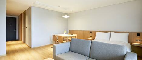 Ein modernes Hotelzimmer mit grauem Sofa, einem Holztisch, einem Bett mit weißer Bettwäsche und einem kleinen Schreibtisch mit Lampe.