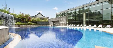 Una piscina con borde de azulejos, una cascada y un edificio moderno con ventanales grandes al fondo.