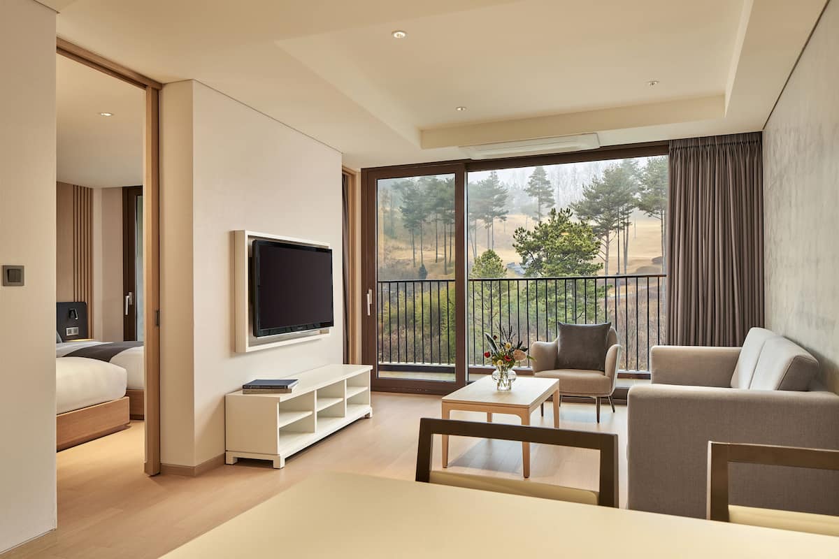 A modern hotel room with a large balcony, a flat-screen TV, a sofa, a coffee table, and a bed with a wooden headboard.