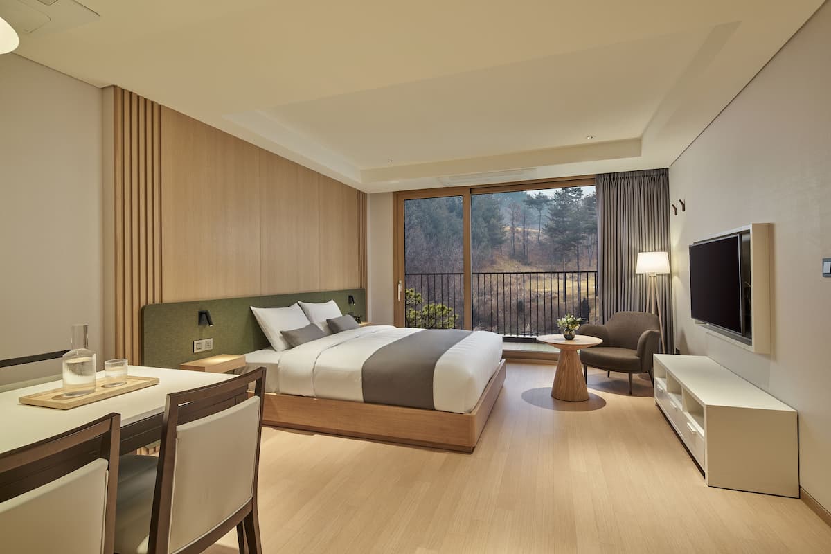 A modern hotel room with a large bed, a desk, a chair, a TV, and a balcony with a view of trees.