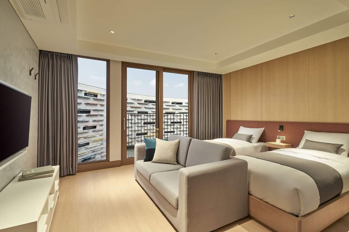 A modern hotel room with a large bed, a sofa, a TV, and a view of a building with balconies.