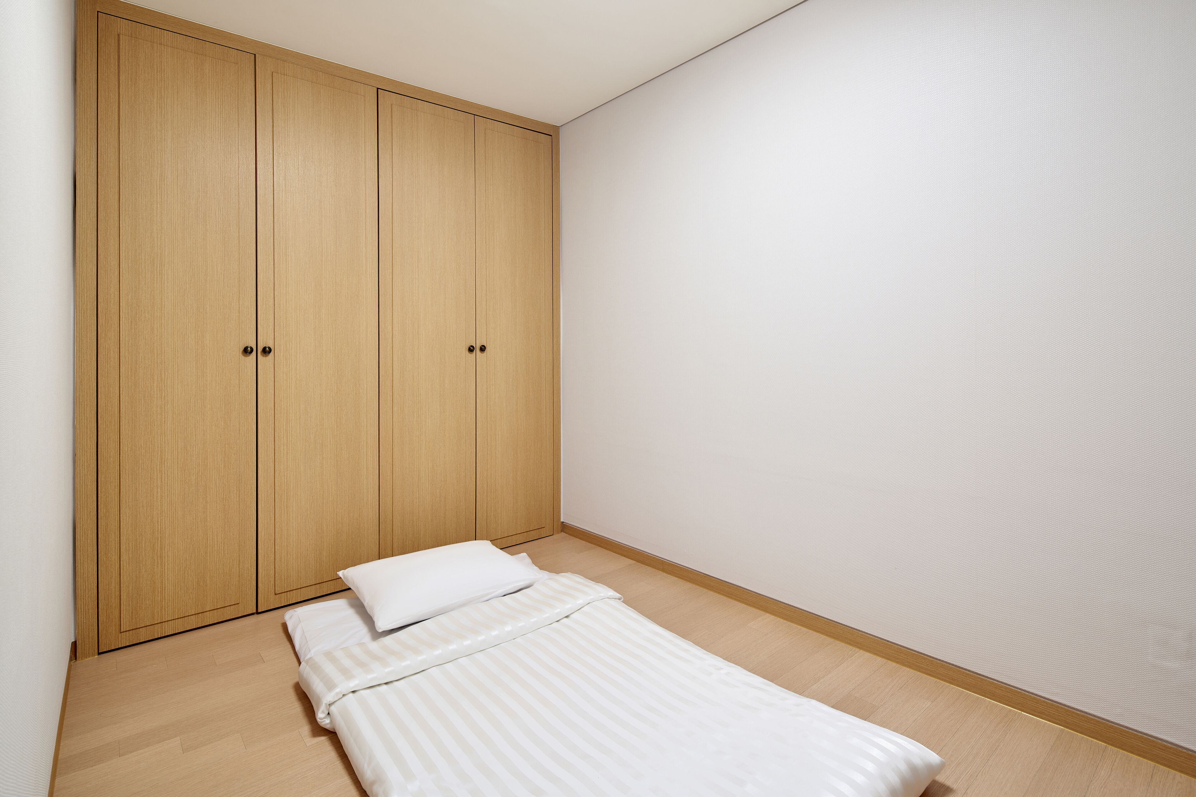 Quarto pequeno e bem arrumado com uma cama de solteiro, guarda-roupa de madeira e uma parede sem decoração.