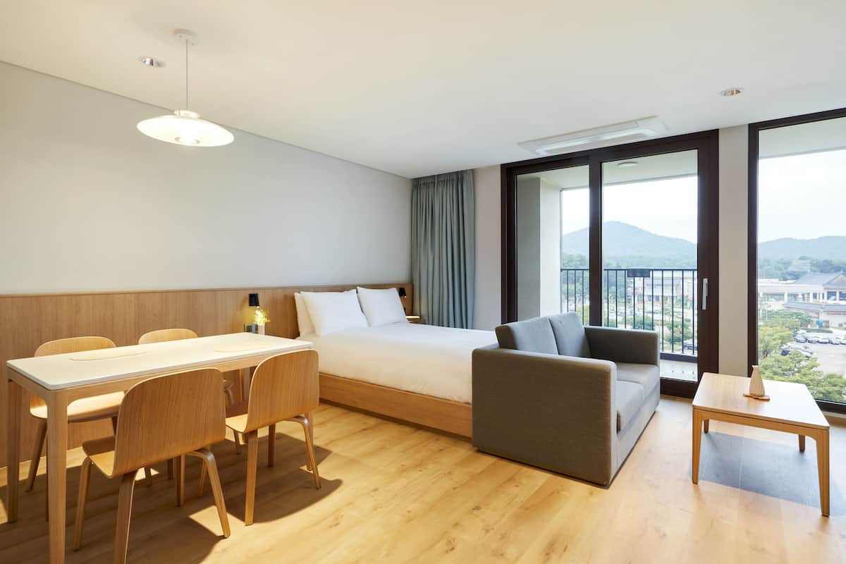 A hotel room with a bed, a sofa, a dining table, and chairs. There is a large window with a view of the outside.