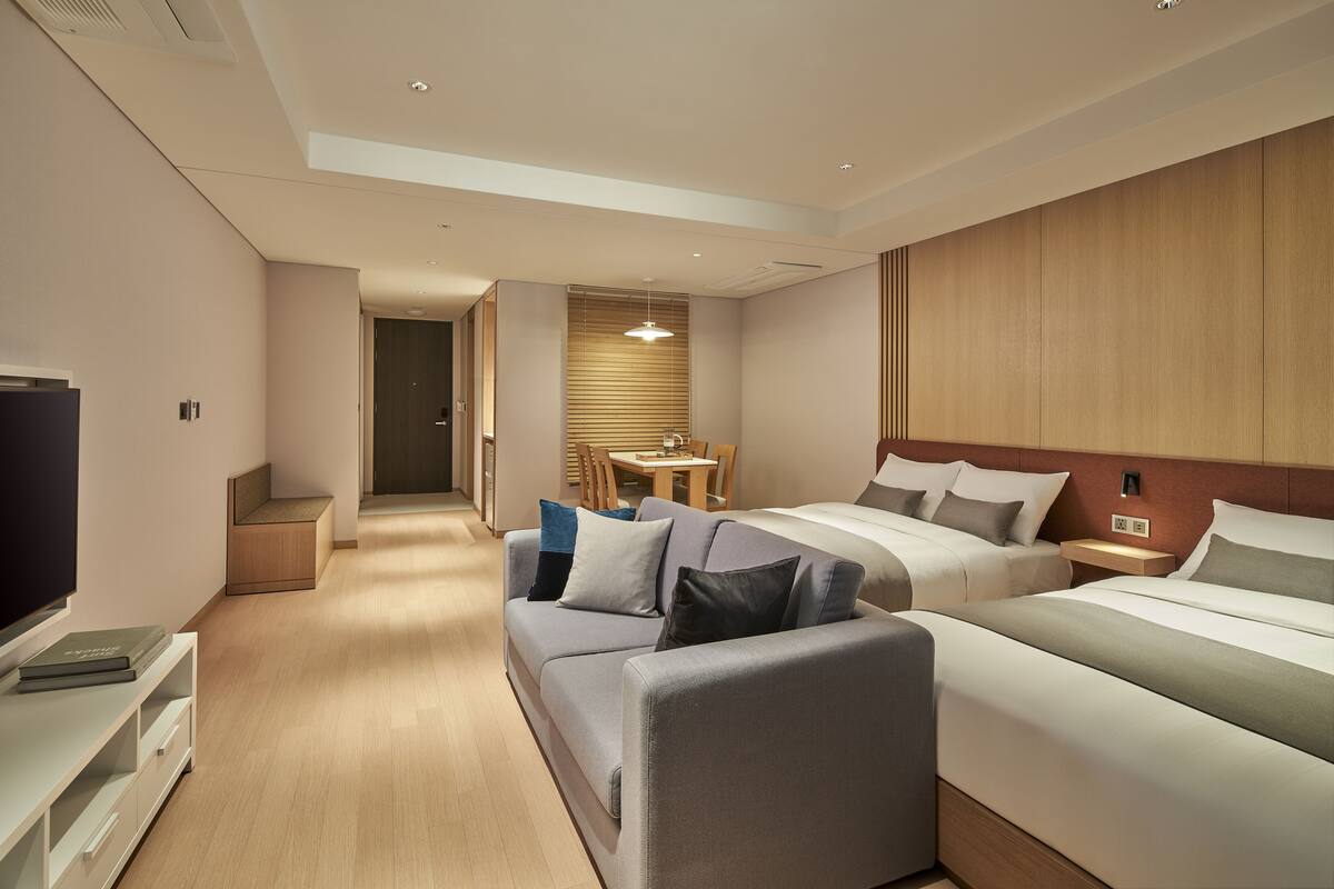 A modern hotel room with a sofa, two beds, a TV, and a small dining area.