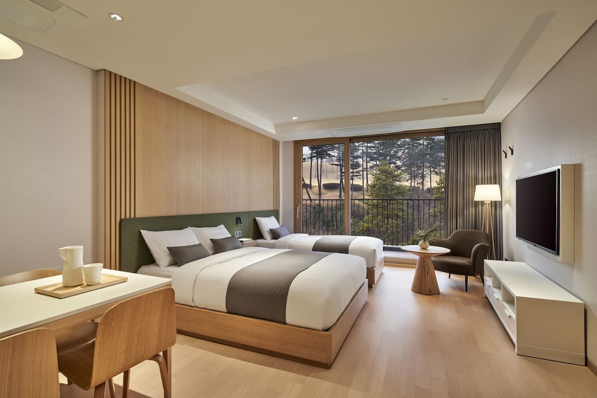 A modern hotel room with a large bed, a desk, a chair, a TV, and a balcony with a view of trees.