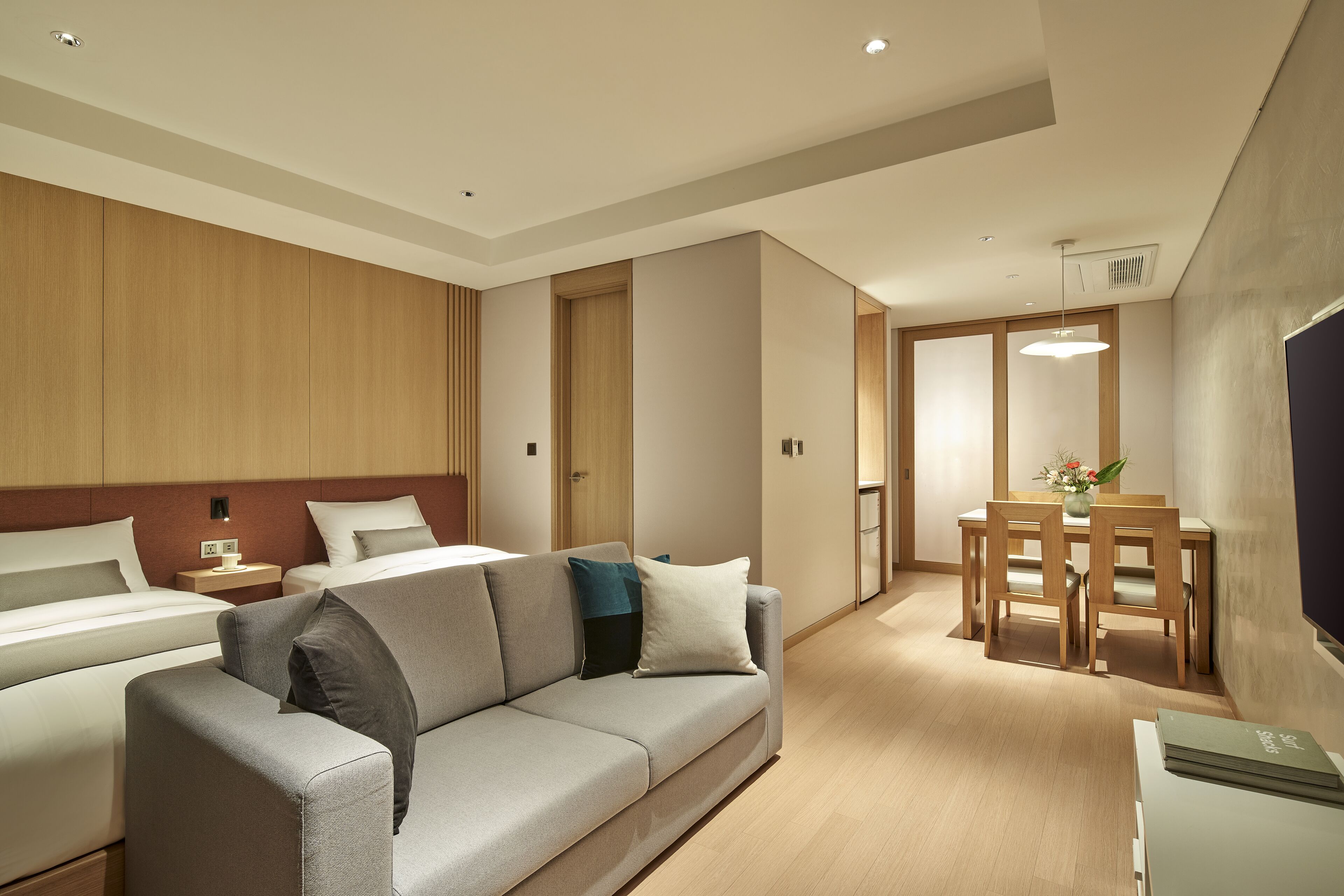 A modern hotel room with a sofa, two beds, a dining table with chairs, and a TV.
