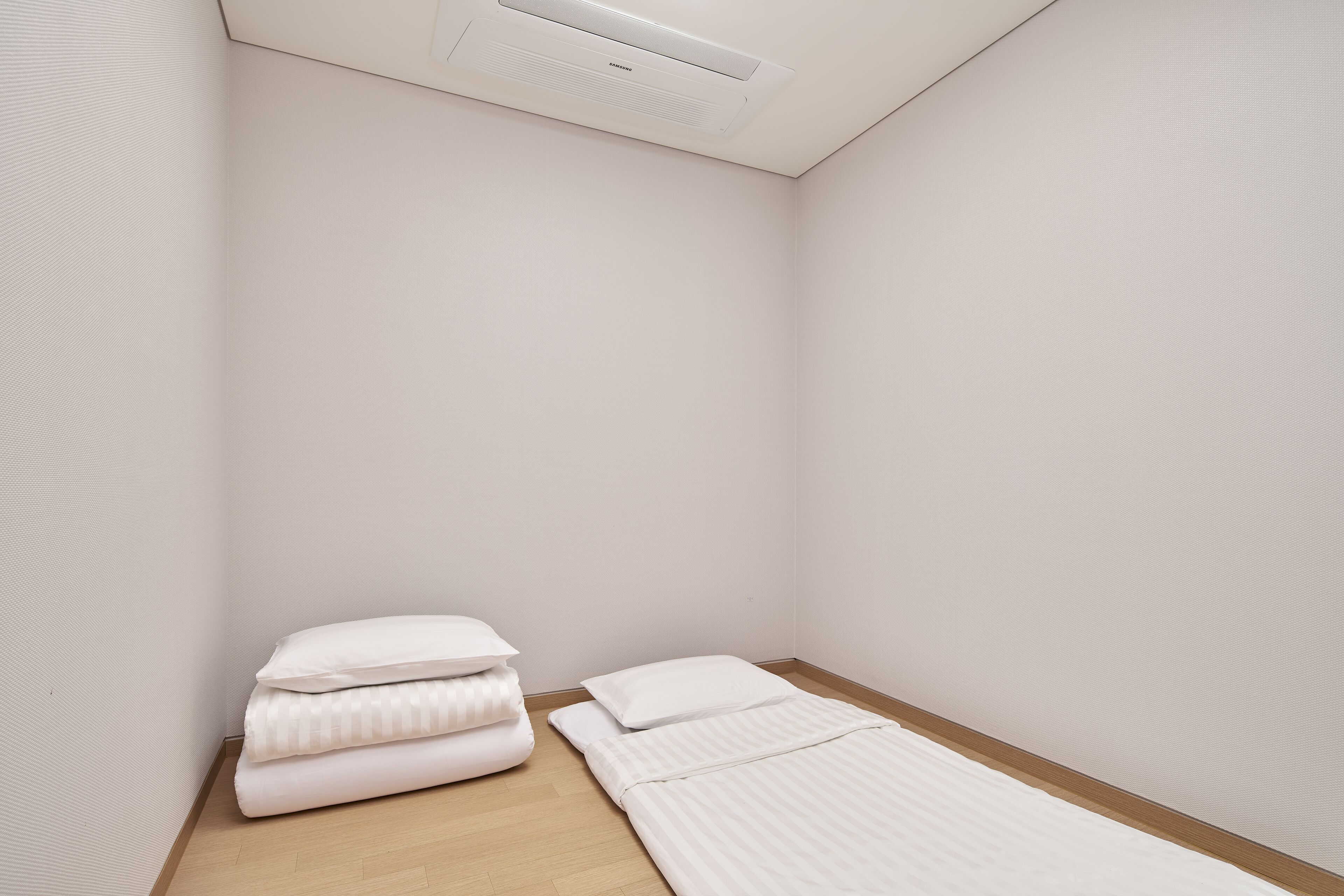 Un petit lit bien fait, avec une literie blanche, dans une chambre d’angle dotée d’un plancher en bois et de murs blancs.