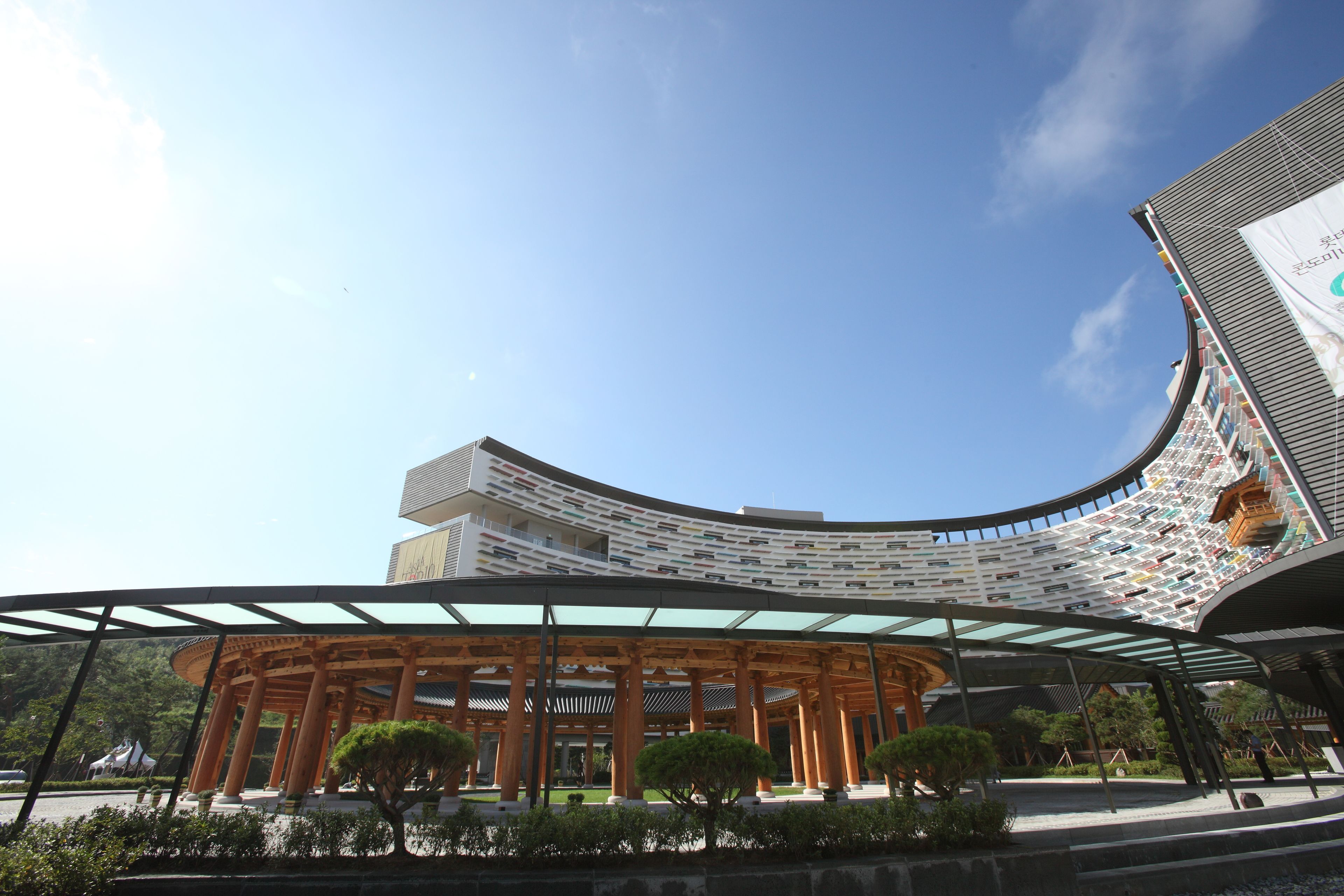 A modern building with a curved facade, a covered pavilion with wooden columns, and a clear blue sky.