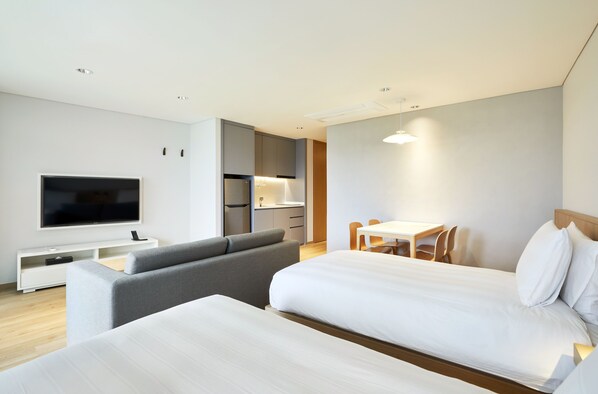 A modern hotel room with a large bed, a sofa, a TV, a dining area, and a kitchenette.