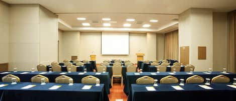 Un salón de conferencias con varias mesas, sillas y una pantalla para proyector.