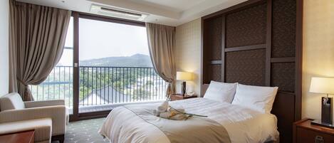 Habitación de hotel con una cama grande, una zona de estar con una silla, una mesita de noche con lámpara y vistas al exterior desde un balcón.