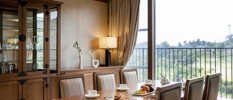 A dining area with a wooden table, white chairs, and a view of a green landscape through large windows.