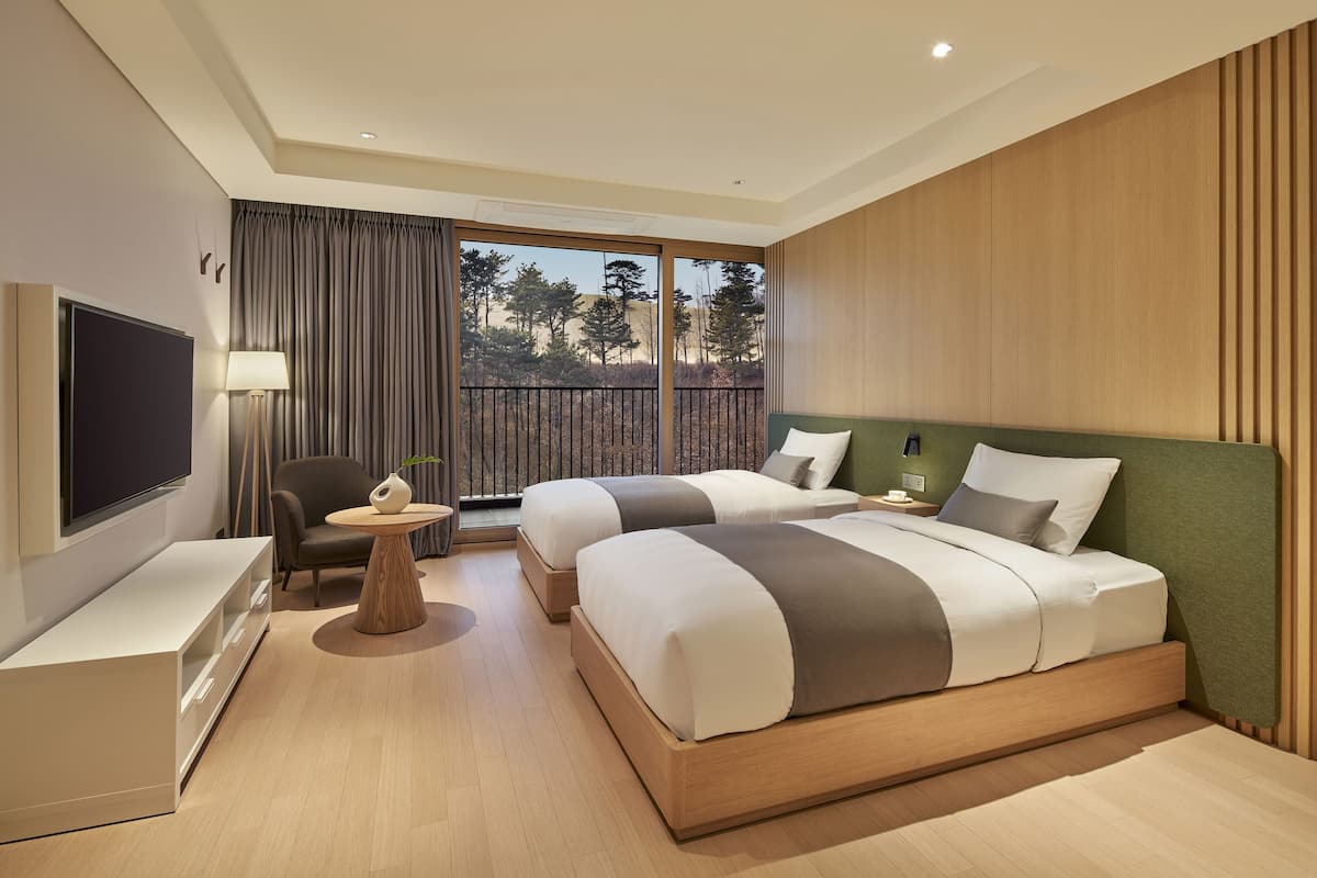 A modern hotel room with two beds, a TV, a desk, and a balcony with a view of trees.