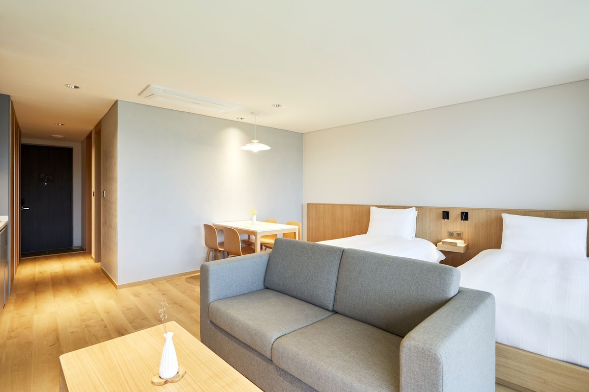 A modern hotel room with a grey sofa, a wooden bedside table, a bed with white linens, and a small desk with a chair.