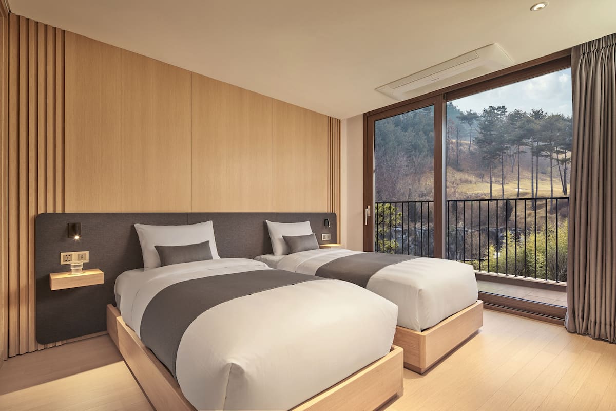 A modern bedroom with two beds, a nightstand, and a large window with a view of trees.
