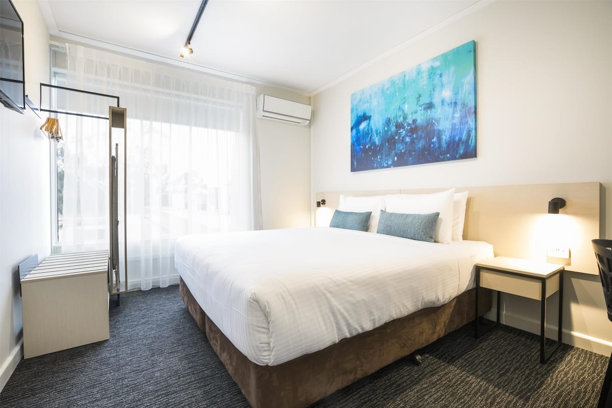 A hotel room with a large bed, bedside table, and a window with sheer curtains.