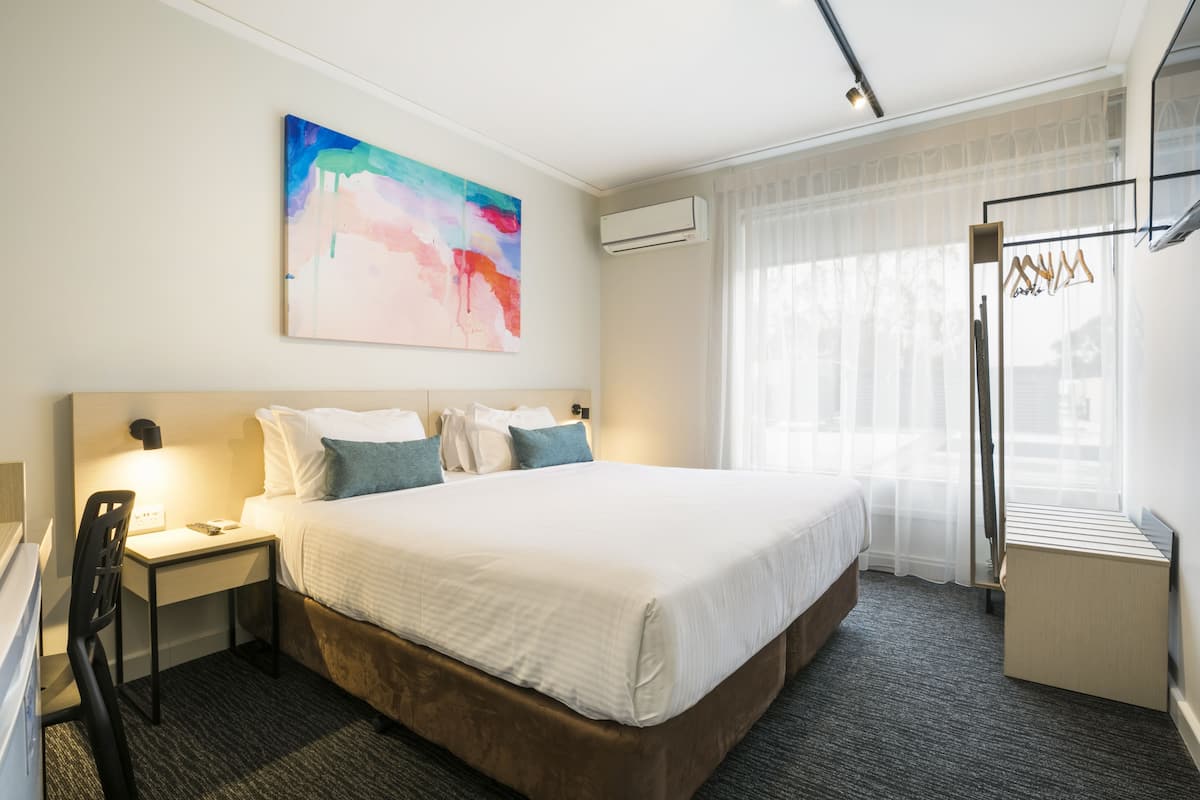 A hotel room with a large bed, a desk with a chair, a bedside table, an air conditioning unit, a window with sheer curtains, and a colorful abstract painting on the wall.