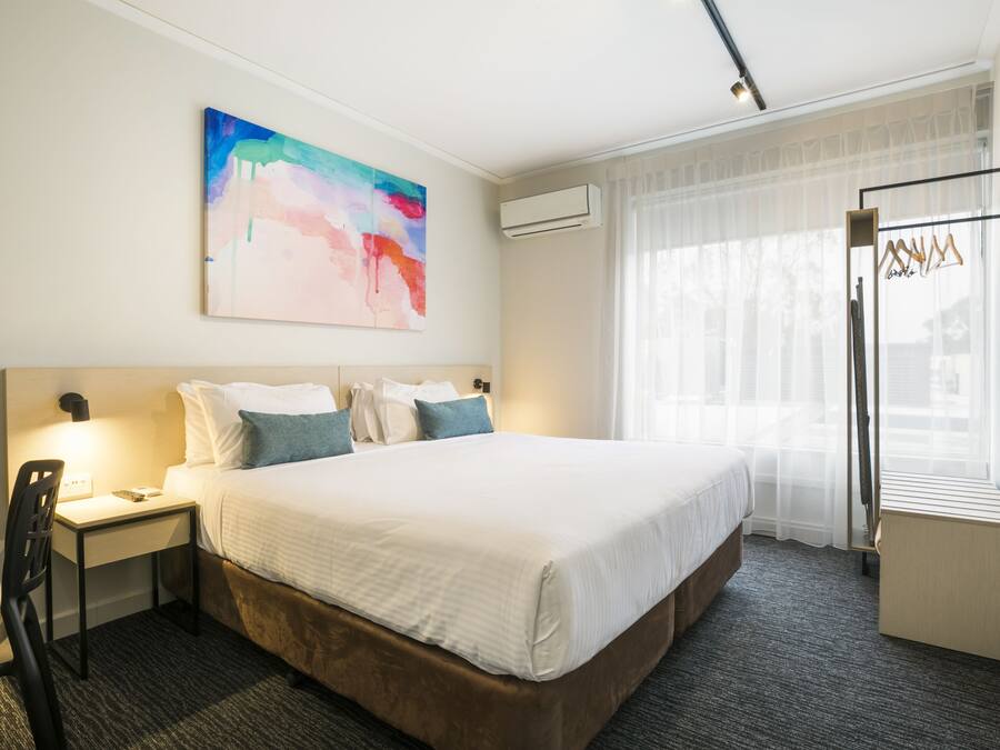 A hotel room with a large bed, a desk with a chair, a bedside table, an air conditioning unit, a window with sheer curtains, and a colorful abstract painting on the wall.