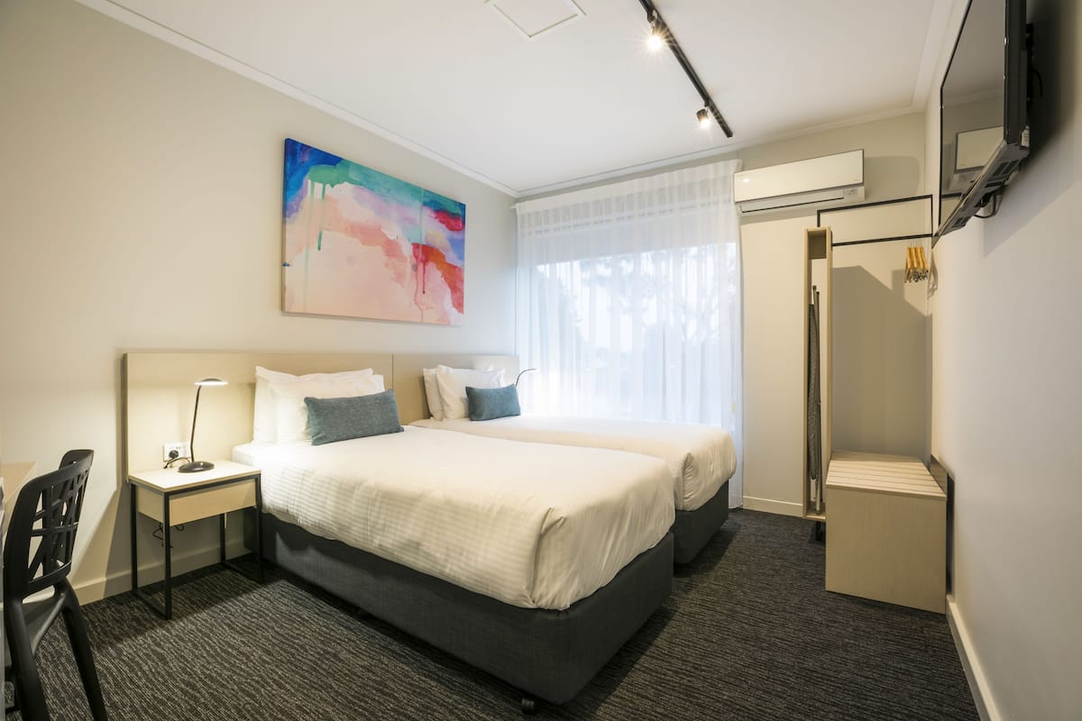 A hotel room with two beds, a desk, a chair, a painting on the wall, and a shower area.