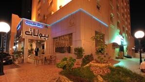 Exterior de un hotel de noche con señalización iluminada, mobiliario exterior y plantas en macetas.