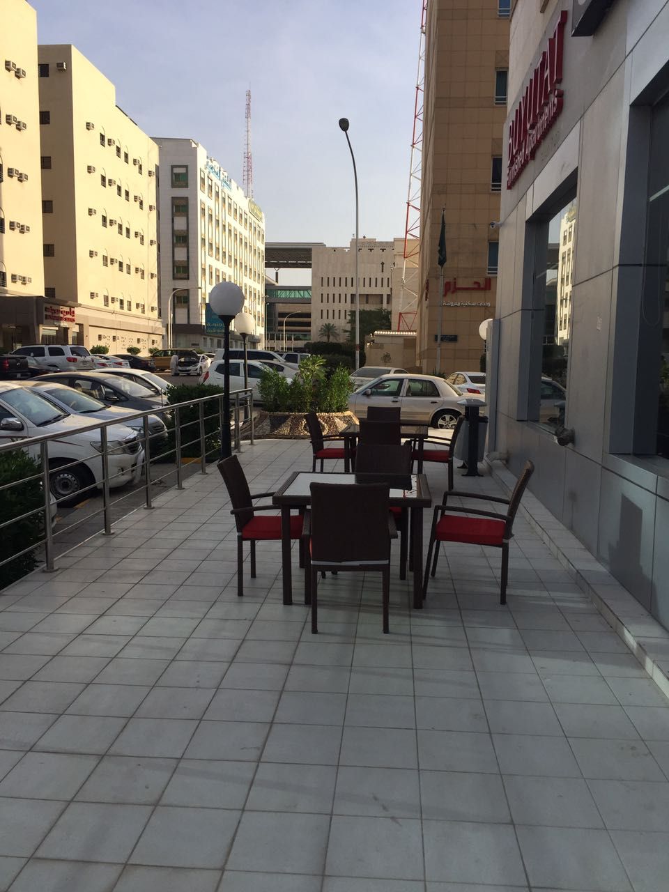 An outdoor seating area with tables and chairs, a parking lot with cars, and buildings in the background.