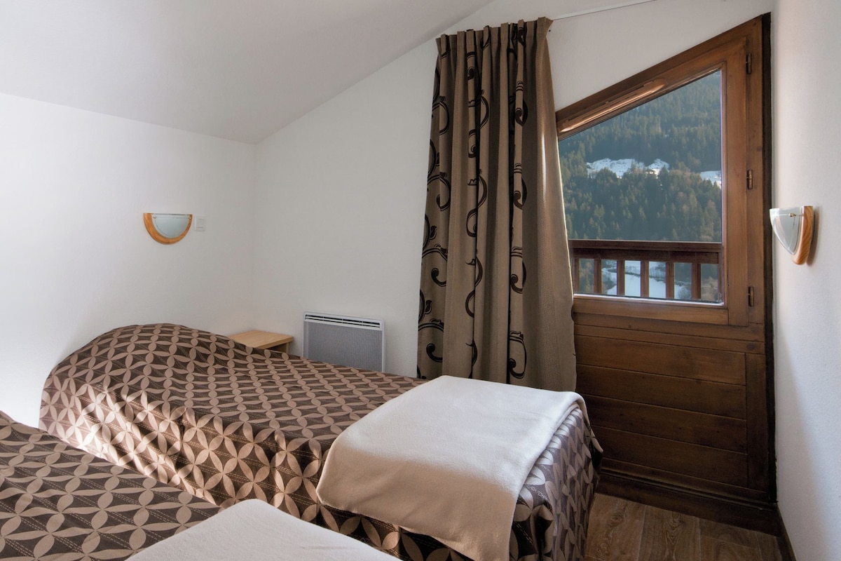 Une chambre avec deux lits, une fenêtre donnant sur une forêt, une porte en bois et un luminaire fixé au mur.