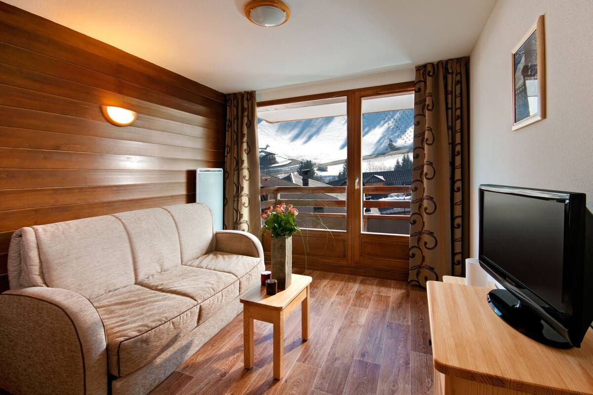 Een woonkamer met een houten vloer, een beige bank, een kleine houten tafel met een bloemenvaas, een flatscreen televisie op een houten standaard, en een groot raam met gordijnen.