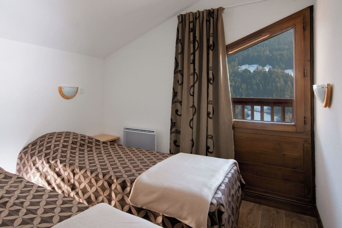 A bedroom with two beds, a window with a view of a forest, a wooden door, and a wall-mounted light fixture.
