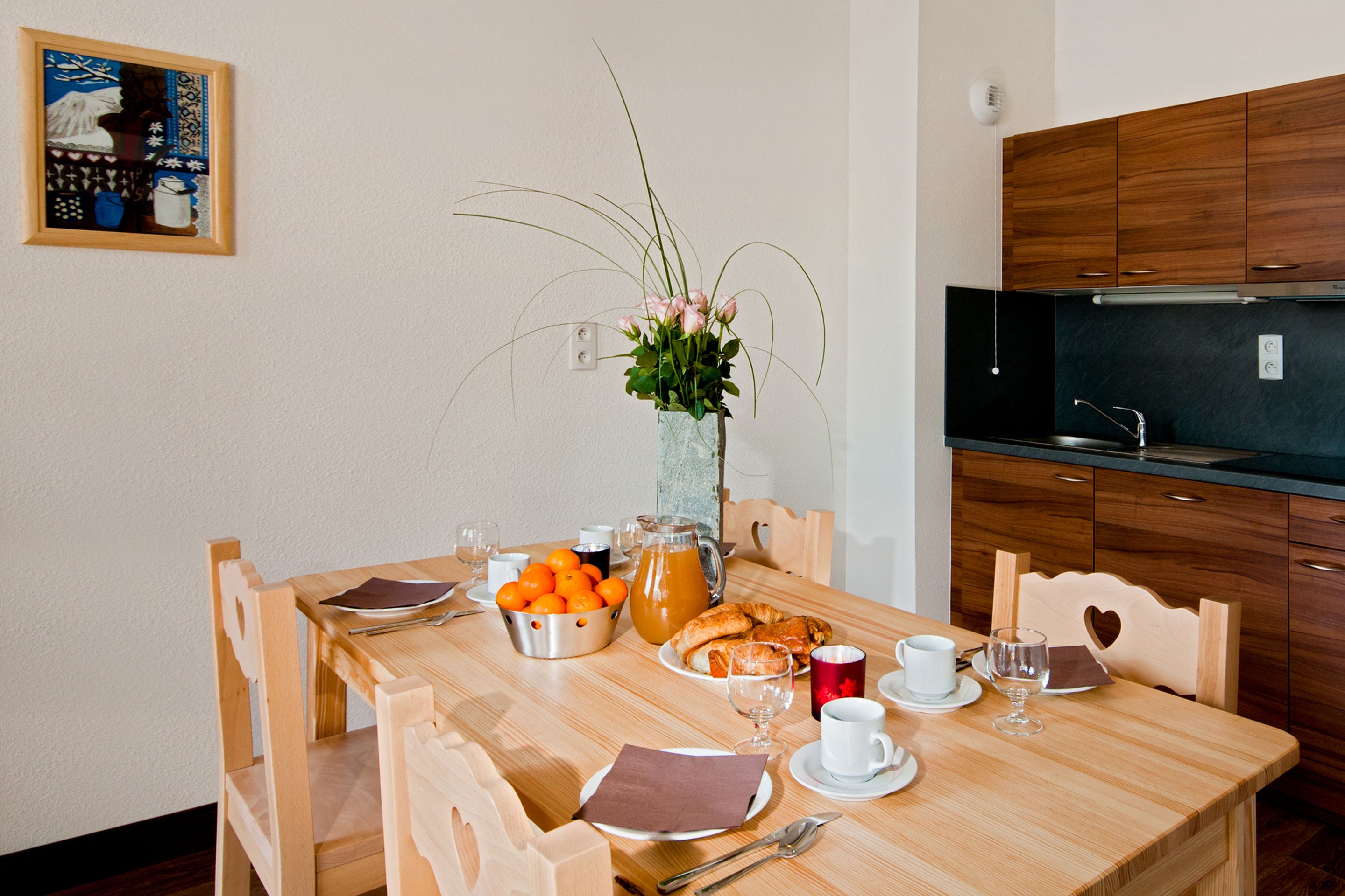A dining table set with breakfast items, a vase with flowers, and wooden chairs.