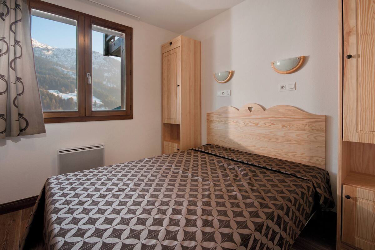 Een slaapkamer met een bed, een raam met uitzicht op besneeuwde bergen, een houten garderobe en een nachtkastje met spiegel.