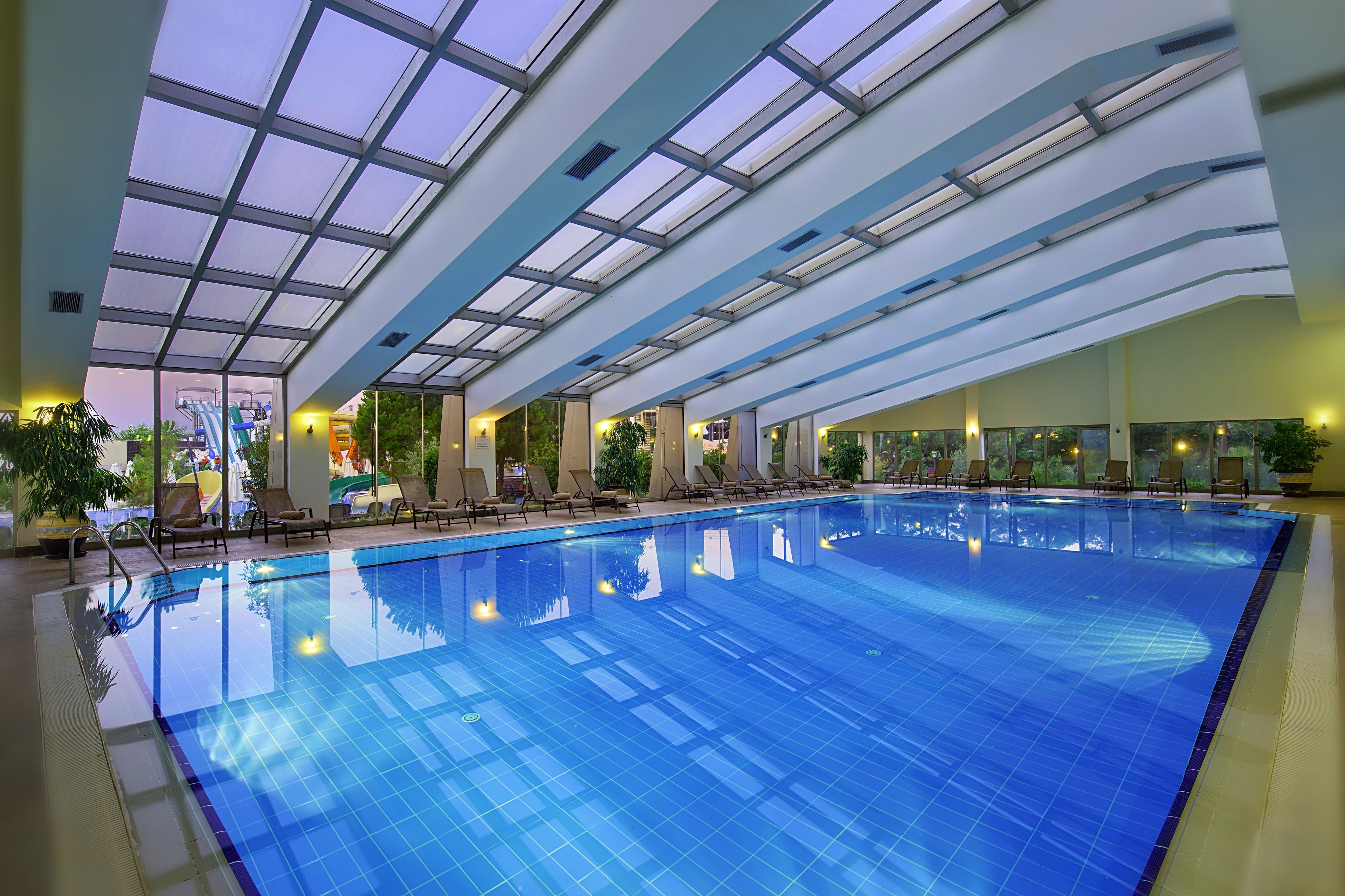 An indoor swimming pool with clear glass ceilings, lounge chairs, and potted plants.