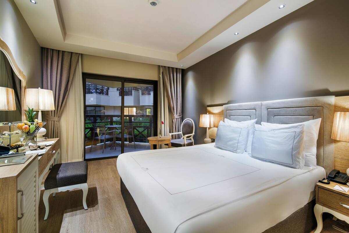 Ein Hotelzimmer mit einem großen Bett, Nachttischen, einem Schreibtisch mit Spiegel und einem Balkon mit Blick auf die Stadt.