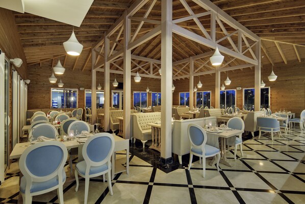 A restaurant with a high ceiling, wooden beams, and a checkered floor. Tables and chairs are set for dining. - Bellis Deluxe Hotel (Serik)
