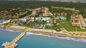 Eine Luftaufnahme eines Küstenresorts mit mehreren Gebäuden, einer Pier, die ins Meer ragt, und einem Strandbereich mit Liegestühlen.